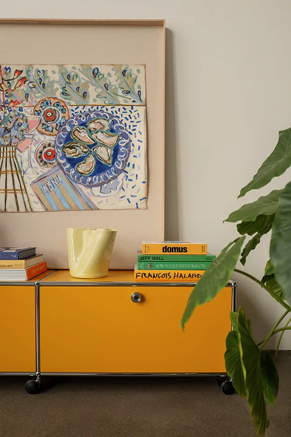 Goldgelbes USM Haller Lowboard mit Klapptüren & Rollen. Darauf Lampe, Vase & Bücher. Rechts daneben steht ein Fahrrad.